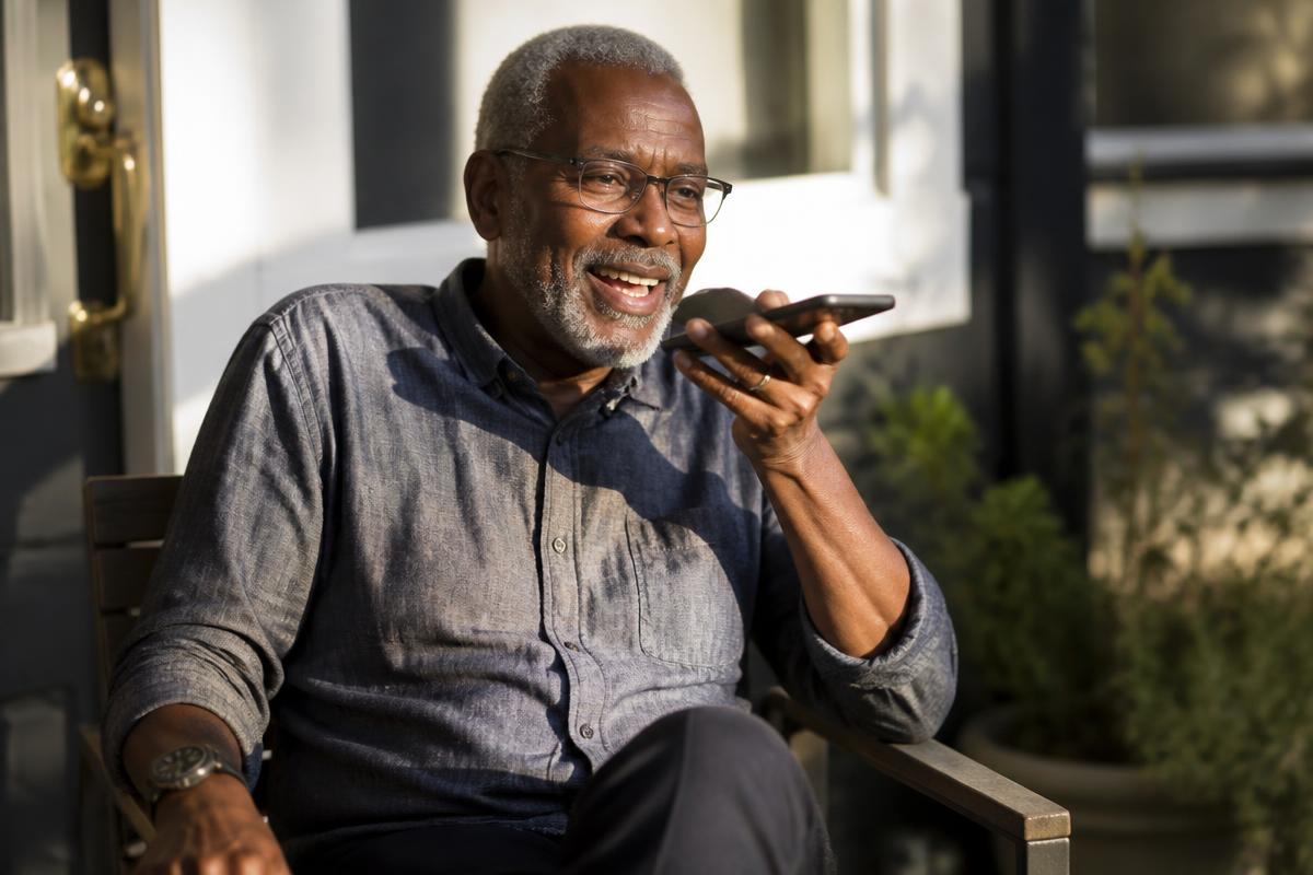 Older man speaking to his smartphone on a sunny porch.